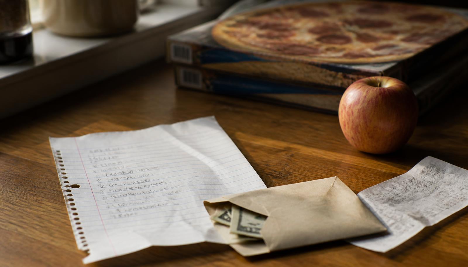 A child's handwritten grocery list beside an envelope of cash, a receipt, an apple, and two frozen pizza boxes on a kitchen counter